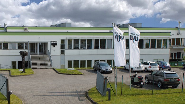 Ein weißes Flachdachgebäude mit grünen Streifen an der Oberkante, wehenden Fahnen, parkenden Autos und einem offenen Einfahrtstor.