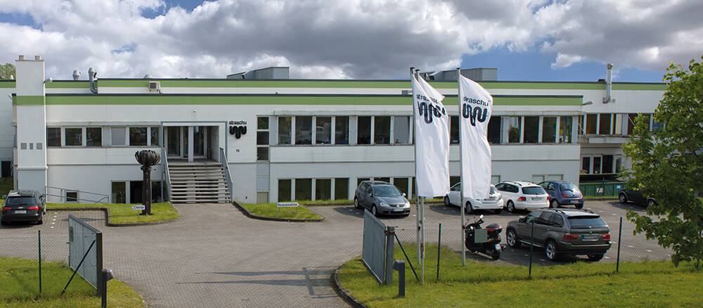 Ein weißes Flachdachgebäude mit grünen Streifen an der Oberkante, wehenden Fahnen, parkenden Autos und einem offenen Einfahrtstor.