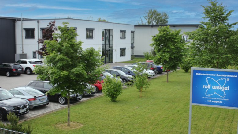 Ein Gebäude mit parkenden Autos davor, Begrünung und ein Firmenschild von rolf Weigel.