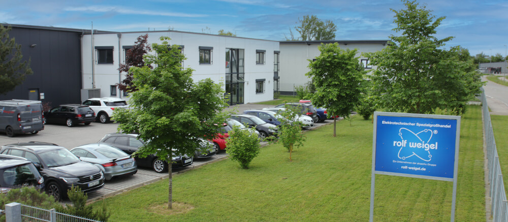 Ein Gebäude mit parkenden Autos davor, Begrünung und ein Firmenschild von rolf Weigel.