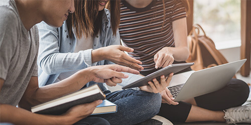 Drei Schüler sitzen im Schneidersitz auf dem Boden und diskutieren mit Büchern, einem Tablet und einem Notebook in den Händen.