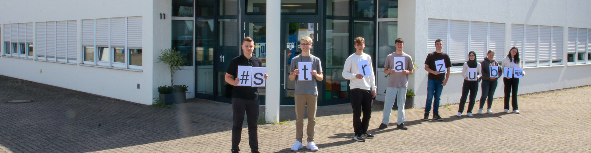 2024_08_Slider_Azubibegrüßung_2 Junge neue Auszubildende stehen in einer Reihe und halten die Buchstaben für das Wort "strazubi"