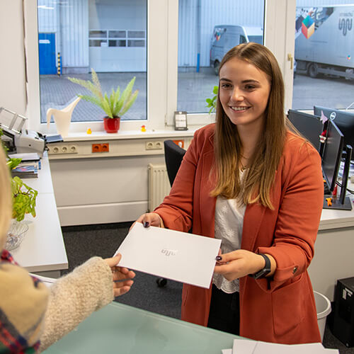 Eine junge Frau im orangenen Blazer, die Auszubildende für Büromanagement ist, nimmt ein Papier entegegen.
