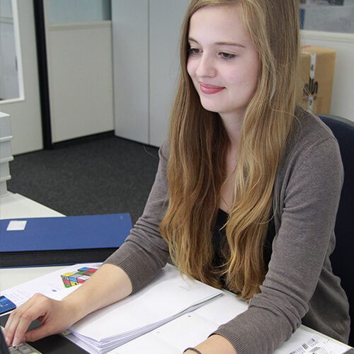 Eine junge blonde Frau, die eine Ausbildung zur Industriekauffrau macht, schaut auf einen Computer.