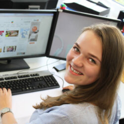 Eine junge Frau sitzt am Computer und schaut nach oben über ihre linke Schulter in die Kamera