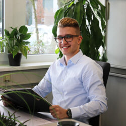 Ein junger Mann mit Brille sitzt am Schreibtisch und hat eine Mappe aufgeschlagen in der Hand