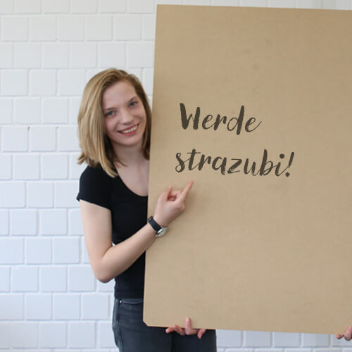 Eine junge blonde Auszubildende hält ein Schild in der Hand, auf dem ``Werde strazubi!`` steht.