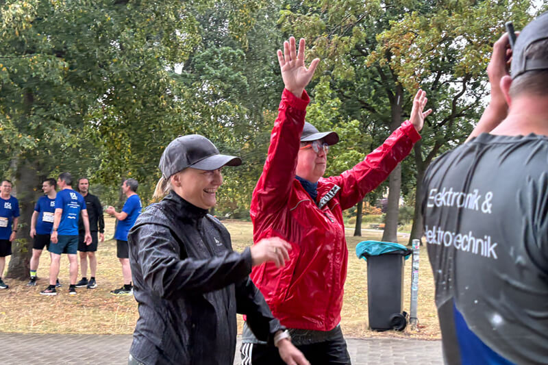 Zwei Frauen laufen beim Firmenlauf, die Frau rechts in einer roten Jacke hebt ihre Arme hoch.