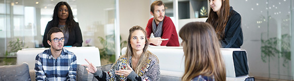 Junge Leute sitzen und stehen in einer Diskussionsrunde in einem Büro auf weißen Sofas