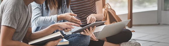 Drei Schüler sitzen auf dem Boden mit Laptop, Tablet und Büchern. Sie zeigen auf ein Tablet, das ein Mädchen in der MItte hält.